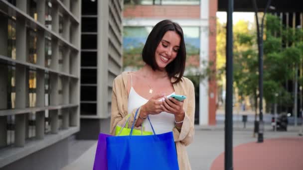 Junge erwachsene fröhliche Frau kauft mit dem Handy online über eine Marktplatz-App ein. Lächelnde Frauen, die Einkaufstüten tragen und Spaß daran haben, einen Verkauf auf dem Handy zu verfolgen. Glückliche kaukasische Dame. Hoch - Filmmaterial, Video
