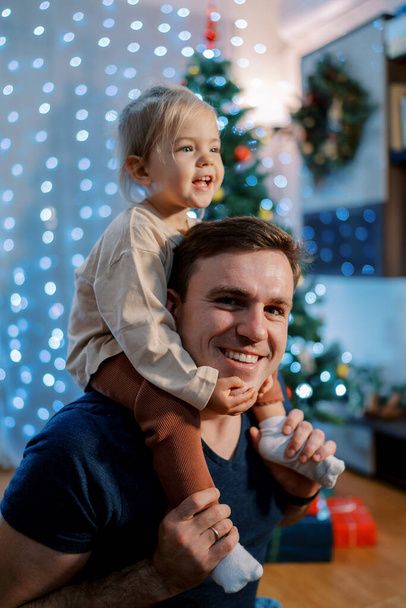 Niña sonriente sosteniendo la barbilla de su padre mientras se sienta sobre sus hombros cerca de un brillante árbol de Navidad. Foto de alta calidad - Foto, Imagen