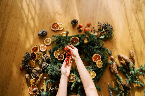 Weibliche Hände halten einen Zweig roter Beeren über einen auf dem Boden liegenden Weihnachtskranz. Ansicht von oben. Hochwertiges Foto - Foto, Bild