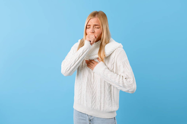 Huolestunut nuori nainen valkoisessa neulotussa puserossa, kädet suun lähellä, yskä tai huonovointisuus kirkkaan sinistä taustaa vasten - Valokuva, kuva