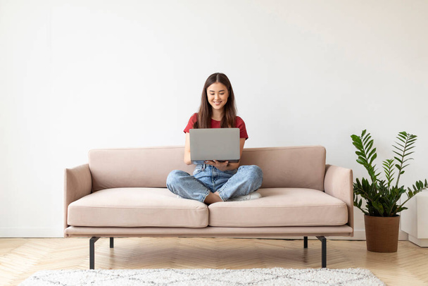 Portret pięknej młodej Azjatki korzystającej z laptopa podczas siedzenia na kanapie w domu, uśmiechnięta Tysiącletnia kobieta pisząca na komputerze, zakupy lub badania online we wnętrzu salonu, kopia miejsca - Zdjęcie, obraz