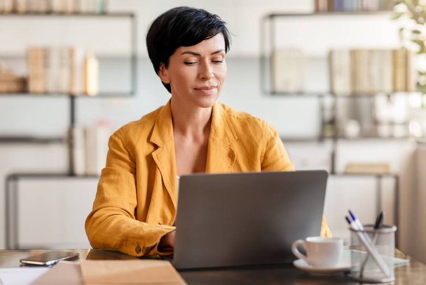 Geschäftsfrau mittleren Alters arbeitet am Laptop und surft im Internet, sitzt am Schreibtisch in modernen Büroräumen. Online-Karriere- und Unternehmerkonzept - Foto, Bild