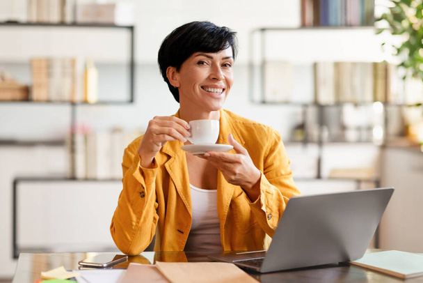 Contented middle aged business lady with brunette haircut enjoying tea at workplace desk, smiling at camera, sitting in modern office interior. Balancing career and lifestyle - Photo, Image