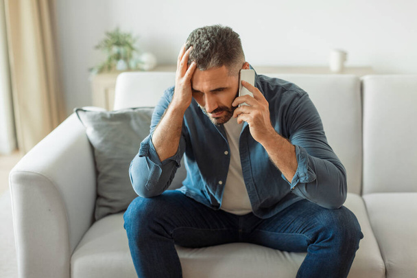 Középkorú európai férfi vesz részt nehéz beszélgetés a mobiltelefon, megható fej frusztrált kifejezés, amelynek problémái kommunikálni telefonon ül a kanapén otthon - Fotó, kép