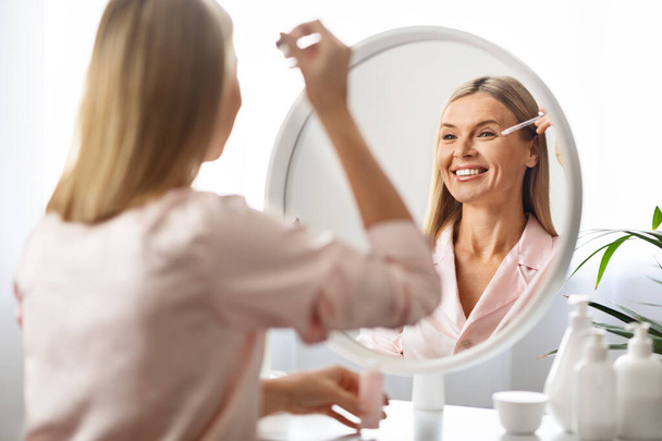 Anti-Aging Cosmetics. Beautiful Mature Female Applying Face Serum Near Mirror, Happy Middle Aged Woman Making Daily Skincare Routine At Home, Testing New Products, Selective Focus On Reflection - 写真・画像