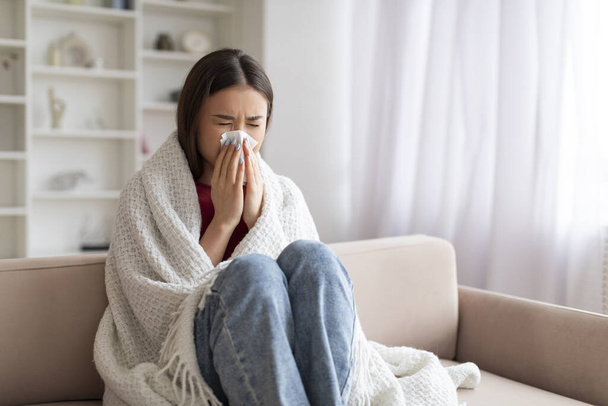 Молодая больная азиатка, сморкающаяся в бумажную ткань, сидя дома на диване, покрытая одеялом, портрет больной кореянки, страдающей насморком, чувствующей себя нездоровой, имеющей сезонный грипп - Фото, изображение