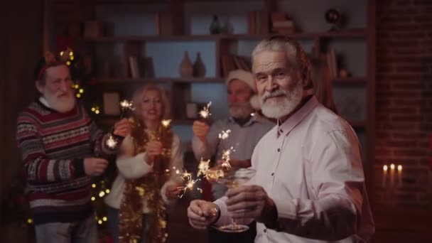 Portret przystojnego starszego pana stojącego, trzymającego kieliszek iskrzącego wina, podczas gdy jego przyjaciele tańczą na tle. Przyjęcie noworoczne. Świętujmy razem, ciesząc się, tańcząc i śmiejąc. Nastrój świąteczny - Materiał filmowy, wideo