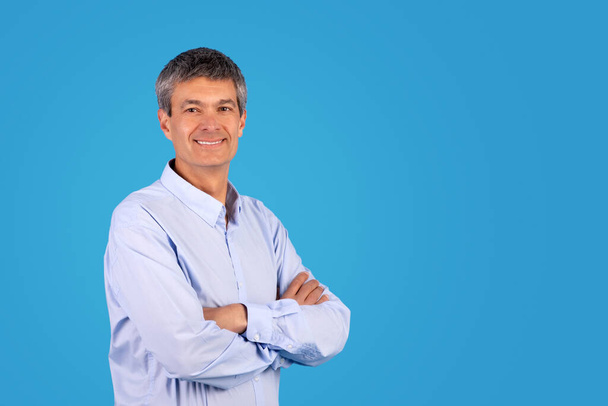 Positiv reifer Mann in lässigem blauem Hemd posiert selbstbewusst vor Studiokulisse, überkreuzt im Stehen die Hände, wirbt lächelnd für Angebot, freie Fläche für Werbung - Foto, Bild