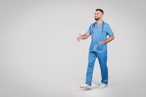 Mladý evropský lékař v modré uniformě sebevědomě kráčí po šedém pozadí, ztělesňuje profesionální lékařskou koncepci, v celé délce, volný prostor - Fotografie, Obrázek