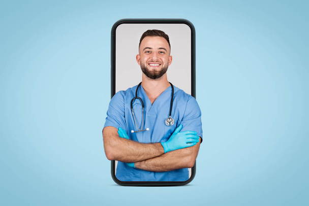 Sebevědomý mladý evropský lékař v modré uniformě, s rukama složenýma s úsměvem na kameru, zobrazený na obrazovce smartphonu na modrém pozadí - Fotografie, Obrázek