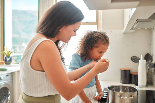 Mixed race mother giving little daughter a wooden spoon to taste. Mother and daughter cooking together in the kitchen. Child tasting flavour in food. - Photo, Image