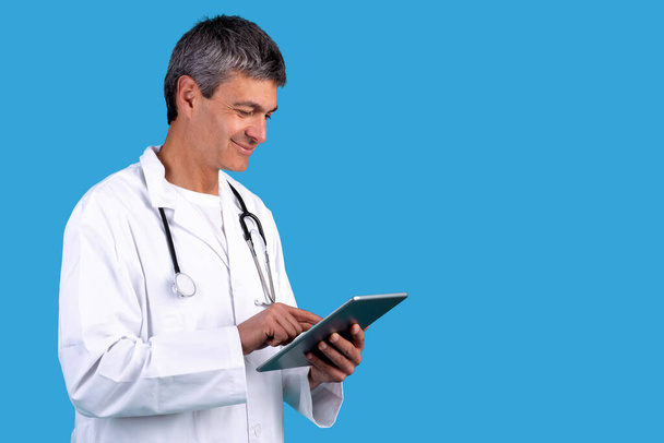 Старший врач веб-серфинга на цифровой планшет позирует в белом мундире на синем фоне студии. Здравоохранение и цифровые технологии для современных медицинских обследований. Копирование пространства - Фото, изображение