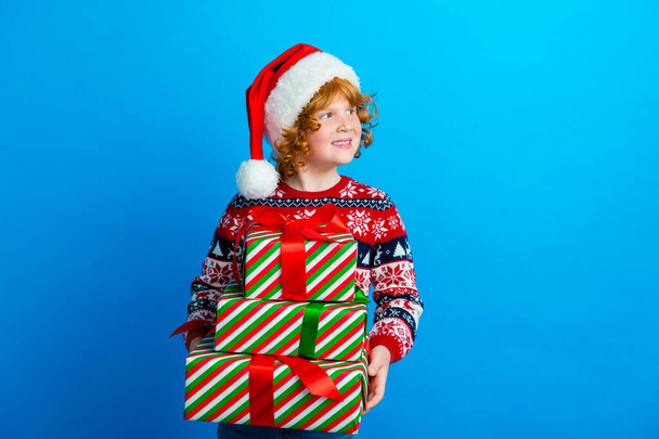 Photo de doux rêve petit garçon porter ornement pull chapeau de Noël tenant des cadeaux à la recherche d'espace vide isolé fond de couleur bleue. - Photo, image