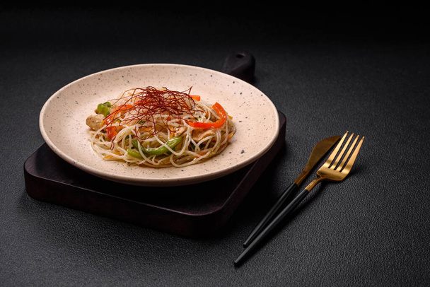 Tasty dish of Asian cuisine with rice noodles, chicken, asparagus, pepper, sesame seeds and soy sauce on dark concrete background - Photo, Image