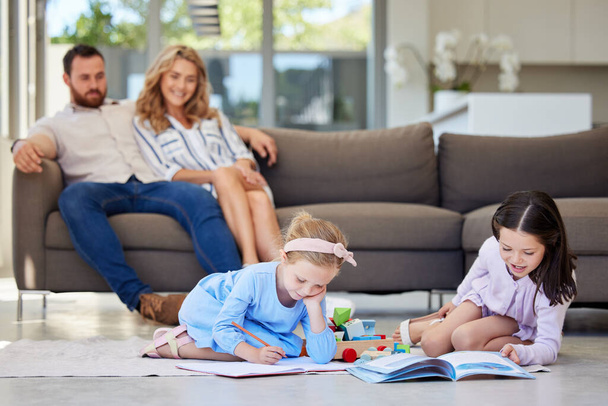 Twee zusjes die samen hun huiswerk maken. Kinderen leren lezen en schrijven liggend op de vloer van de woonkamer met hun ouders kijken op de achtergrond. - Foto, afbeelding