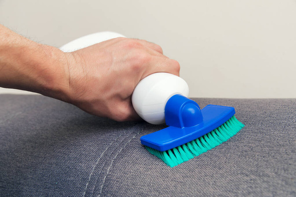 L'homme utilise un produit nettoyant avec une éponge et une brosse pour nettoyer la surface d'un canapé. Nettoyage de la maison. Service de nettoyage et de propreté. - Photo, image