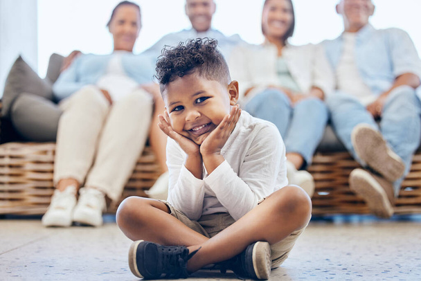 Muotokuva suloinen afrikkalainen amerikkalainen poika istuu jalat ristissä lattialla, kun perhe istuu tuolilla taustalla. Söpö pieni poika hymyilee istuessaan kotona.. - Valokuva, kuva