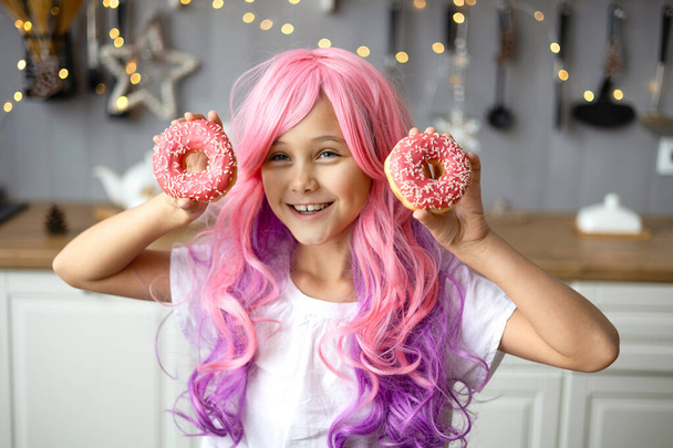 Portrait of a little smiling girl with pink hair and two appetizing donuts in her hands, on a kitchen background. Vanilla Girl. Kawaii vibes. - Photo, Image