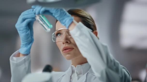 Professional biochemist examining test tube with medicines close up. Focused woman scientist looking at experimental drugs in laboratory. Female chemist in protective glasses making medical research. - Footage, Video
