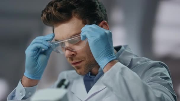 Vědec s bezpečnostními brýlemi zkoumat léky v pokročilé laboratoři zblízka. Vážný muž si nasadil brýle. Důvěryhodný mladý biolog začíná pracovat v biotechnologické laboratoři. - Záběry, video