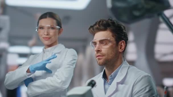 Два колеги з лабораторії посміхаються до камери в передовій клініці крупним планом. Портрет успішних вчених в захисних окулярах в інноваційній лабораторії. Щасливі професійні біохіміки позують у сучасній лікарні - Кадри, відео