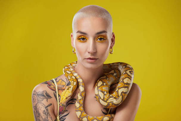 Ύψωσε δυνατά. μια νεαρή γυναίκα που ποζάρει με ένα φίδι γύρω από το λαιμό της σε κίτρινο φόντο - Φωτογραφία, εικόνα
