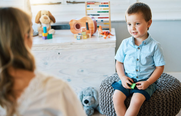 Очаровательный маленький мальчик, сидящий на стуле, разговаривая с кавказским терапевтом. Милый мальчик разговаривает с психологом. Проверить ребенка у психолога в приемной семье перед усыновлением. - Фото, изображение