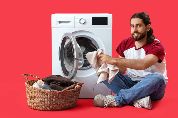 Hombre joven con cesta de lavandería cerca de la lavadora sobre fondo rojo - Foto, Imagen