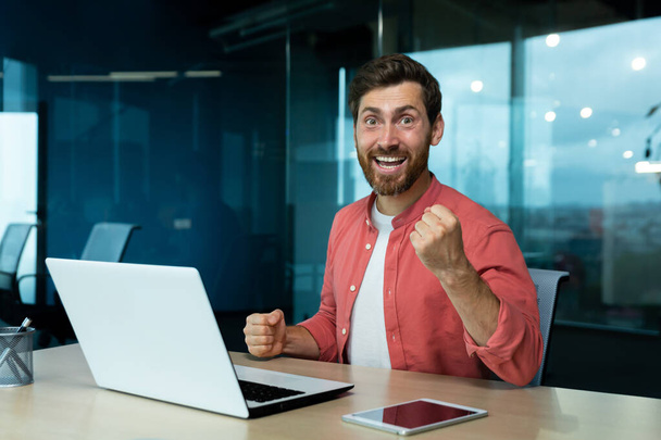 Portrét úspěšného zralého podnikatele v červené košili na pracovišti v kanceláři, muž slaví vítězství a úspěšné výsledky v práci, dívá se do kamery, drží ruku nahoru. - Fotografie, Obrázek
