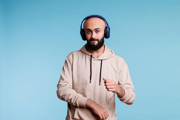 Homme arabe écouter de la musique dans les écouteurs et faire des mouvements avec des mains portrait. Jeune personne détendue portant des écouteurs sans fil dansant tout en appréciant la chanson et regardant la caméra - Photo, image