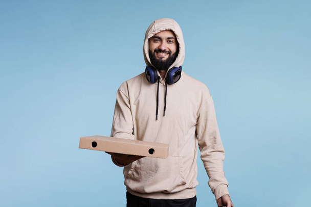 Mensajero árabe sonriente sosteniendo la caja de pizza y mirando a la cámara con expresión alegre. Despreocupado repartidor de pizzería dando comida rápida para llevar estudio retrato sobre fondo azul - Foto, imagen