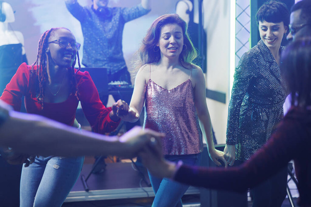 Beztroskie kobiety trzymające się za ręce i tańczące razem podczas imprezy w nocnym klubie. Wesołe młode, zróżnicowane dziewczyny clubbing i zabawy na parkiecie, korzystających z aktywności nocnego życia - Zdjęcie, obraz