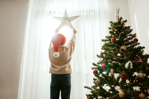 Genç bir kız yeni yıl için evini ve dairesini dekore ediyor. Bir çocuk yılbaşı ağacının yanındaki pencereye yeni yıl yıldızı asar. - Fotoğraf, Görsel