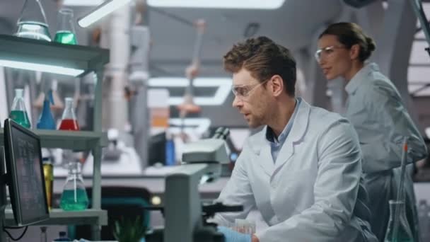 Un par de científicos trabajando juntos en un laboratorio moderno de cerca. Compañeros de laboratorio sonrientes mirando al monitor de computadora haciendo investigación en la clínica. Amistosa mujer química con tableta de consultoría pasante hombre. - Metraje, vídeo