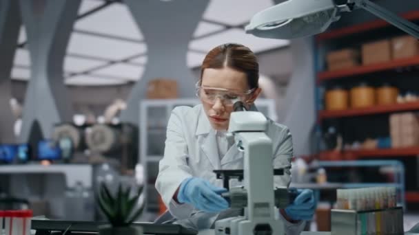 Professional scientist looking in microscope analyzing sample close up. Serious woman biotechnology researcher making notes of experiment results. Female doctor working in pharmacological laboratory. - Footage, Video