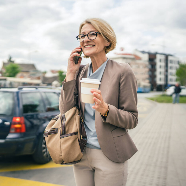 Одна жінка кавказька зріла блондинка з окулярами стоїть на зупинці автобуса вдень використовуйте мобільний телефон, роблячи розмову про дзвінок, чекаючи, поки привід справжня людина скопіює простір пізно для роботи
 - Фото, зображення
