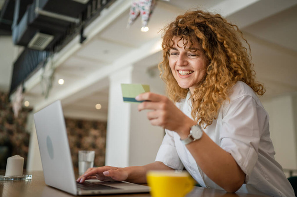 Jedna žena dospělý kavkazský žena s kudrnaté vlasy šťastný úsměv použití kreditní karty nakupování on-line na notebooku v kavárně skuteční lidé e-bankovnictví internet prodej kopírovat prostor pozitivní emoce - Fotografie, Obrázek