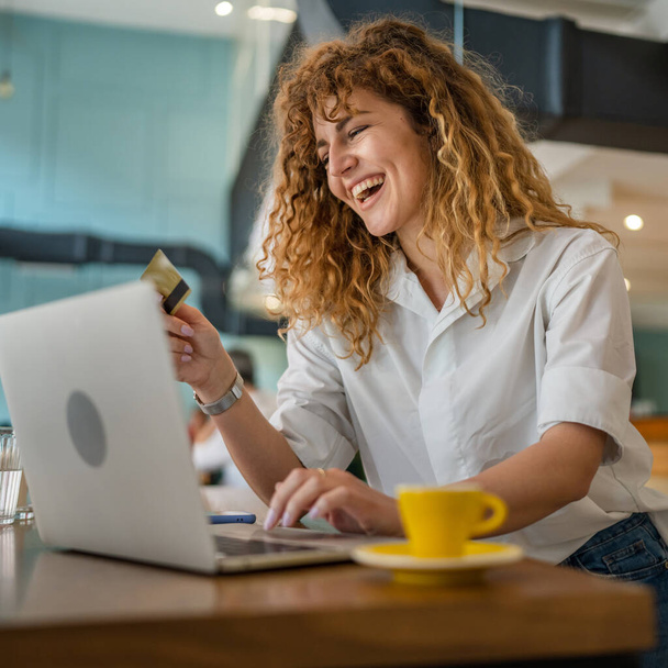 Kıvırcık saçlı, mutlu, yetişkin bir kadın Kafe 'deki laptop bilgisayarından kredi kartı alışverişi yapıyor. Gerçek insanlar e-bankacılık internet indirimi yapıyor. - Fotoğraf, Görsel