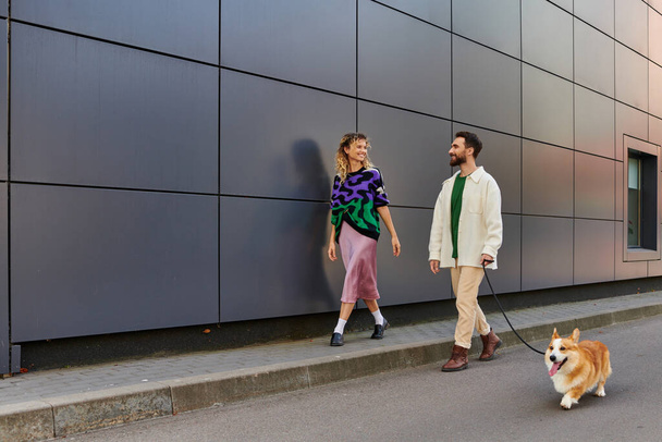 šťastný pár držící se za ruce a procházející se se psem corgi v blízkosti moderní šedé budovy, zvířecí společníci - Fotografie, Obrázek