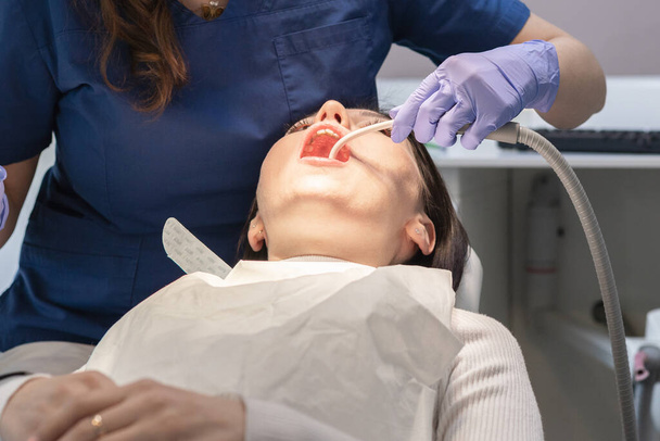 Egy kék egyenruhás és orgonakesztyűs fogorvos eltávolítja a nyálat egy fiatal barna páciens szájába, aki egy széken fekszik, alulról közelről nézve. Koncepció, egészség és orális - Fotó, kép