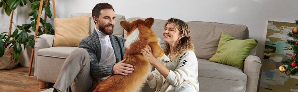 gelukkig paar glimlachen en spelen met schattige corgi hond in modern appartement, gezellige momenten banner - Foto, afbeelding