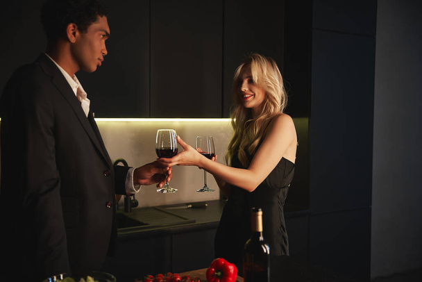 Zarif takım elbiseli yakışıklı Afro-Amerikan adam neşeli kız arkadaşından bir bardak kırmızı şarap alıyor. - Fotoğraf, Görsel