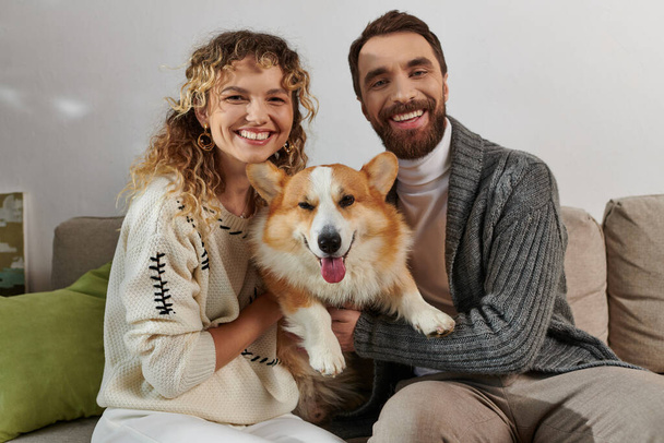 χαρούμενο ζευγάρι σε χειμωνιάτικη ενδυμασία χαμογελώντας και παίζοντας με το σκυλάκι στο μοντέρνο διαμέρισμα, χαρούμενες στιγμές - Φωτογραφία, εικόνα