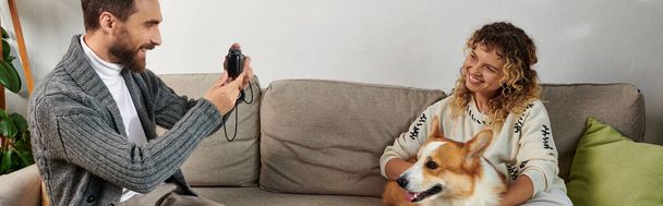 happy man smiling and taking photo of wife with corgi dog on camera in modern apartment, banner - 写真・画像