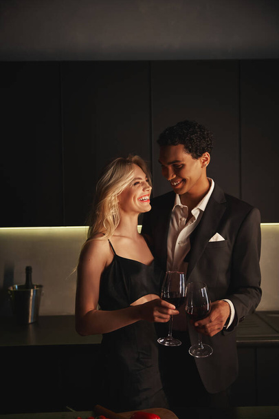 cheerful young multicultural couple in elegant attires enjoying each other and holding wine glasses - Zdjęcie, obraz