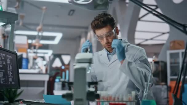 Scientist putting safety glasses working with chemical samples in biochemistry laboratory close up. Focused man chemist holding test tubes opening notepad. Smart doctor making vaccine research in lab. - Footage, Video