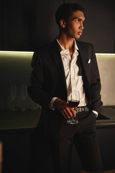 bell'uomo afroamericano dall'aspetto elegante in abito elegante in posa con bicchiere di vino rosso e distogliendo lo sguardo - Foto, immagini