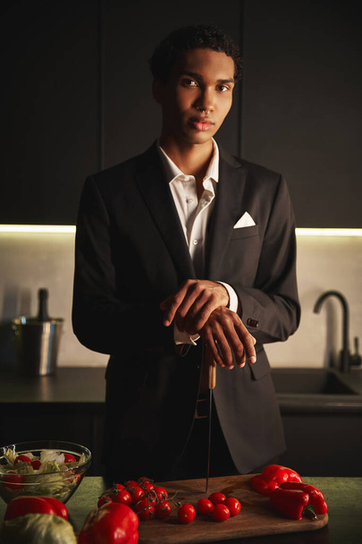 εμφανίσιμος νεαρός άνδρας με κομψό μαύρο κοστούμι ποζάροντας στην κουζίνα του και κοιτάζοντας κατευθείαν την κάμερα - Φωτογραφία, εικόνα