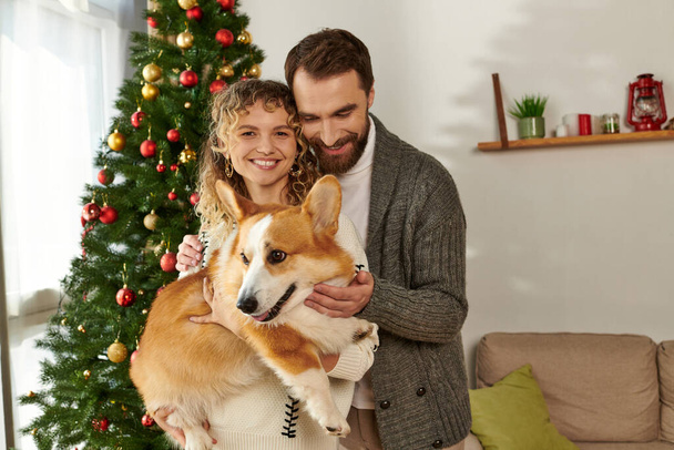 feliz pareja en invierno ropa celebración corgi perro y de pie cerca decorado árbol de Navidad - Foto, imagen