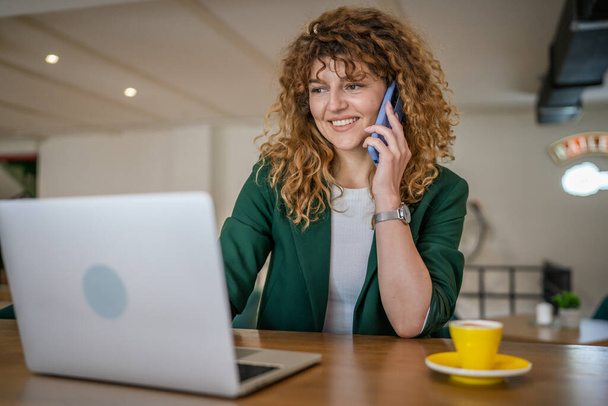 Uma mulher adulta caucasiana fêmea sentar-se no trabalho café no computador portátil fazer uma chamada telefônica falar sorriso feliz confiante sorrindo espaço cópia pessoa real - Foto, Imagem